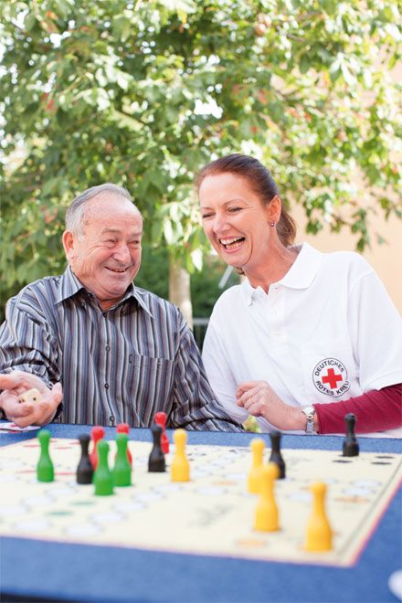 Foto: Eine DRK-Mitarbeiterin spielt 'Mensch ärgere dich nicht' mit einem älteren Herrn. Beide scheinen dabei sichtlich Spaß zu haben und lachen miteinander.